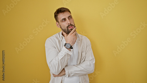 Cool, relaxed yet doubtful expression of a handsome, bearded young hispanic man standing alone against a bold, yellow isolated background