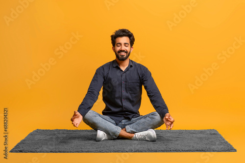 Relaxed young indian man in casual clothes meditating alone, sitting cross-legged on carpe