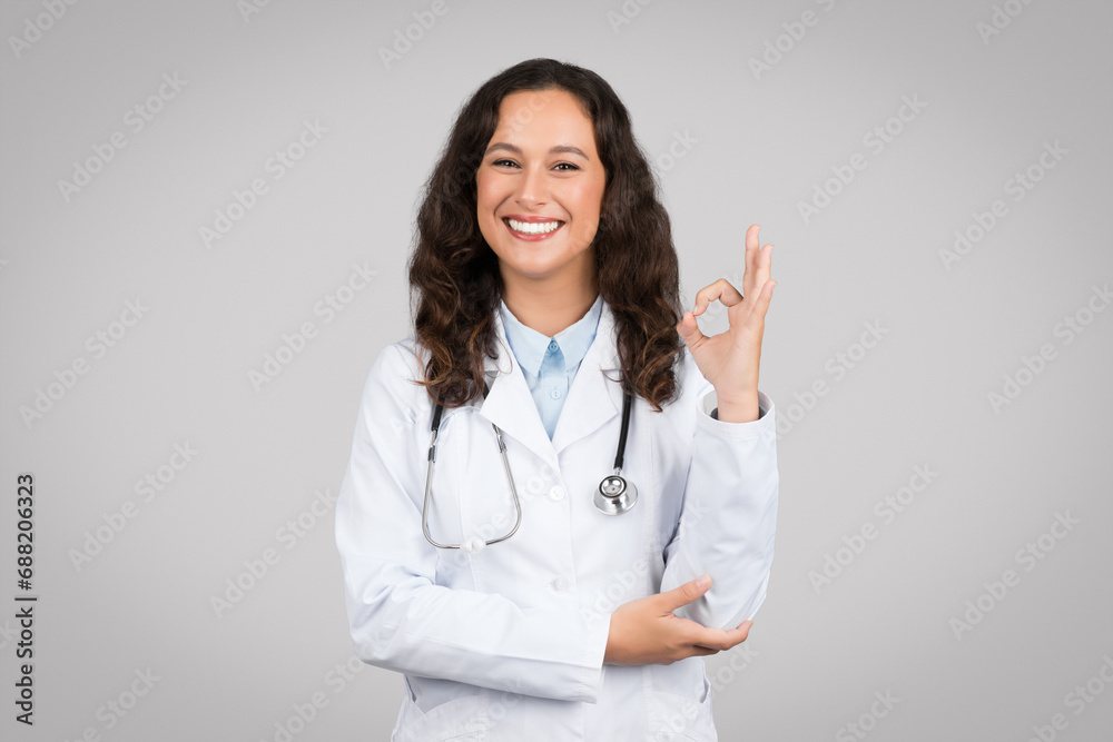 Glad european woman doctor in white coat making ok sign with hand, standing on gray background, smiling at camera Glad european woman doctor in white coat making ok sign with hand, standing on gray background, smiling at camera