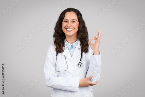 Glad european woman doctor in white coat making ok sign with hand  standing on gray background  smiling at camera