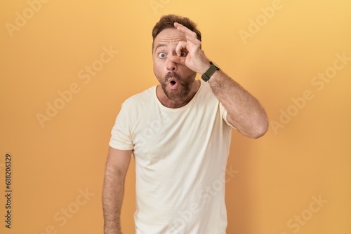 Middle age man with beard standing over yellow background doing ok gesture shocked with surprised face, eye looking through fingers. unbelieving expression.