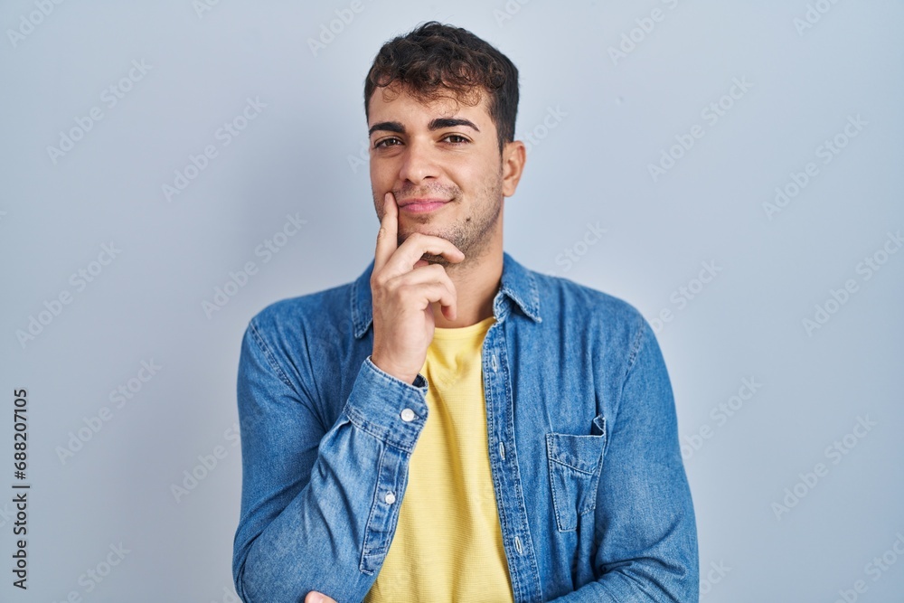 Young hispanic man standing over blue background looking confident at the camera smiling with crossed arms and hand raised on chin. thinking positive. Young hispanic man standing over blue background looking confident at the camera smiling with crossed arms and hand raised on chin. thinking positive.