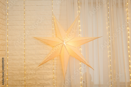Christmas star on golden lights background on wall in living room. Large paper star glows against the backdrop of New Year garland. Atmospheric festive xmas house decorating. Happy 2025 Holidays.