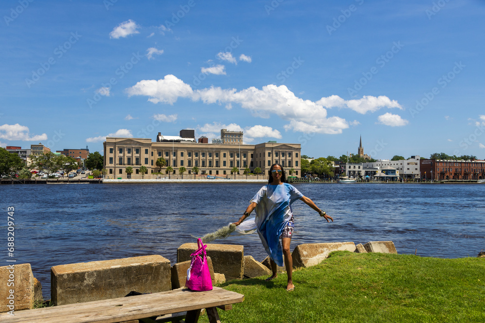 An African American woman with long sisterlocks wearing a blue dress and sunglasses with lush green grass and hotels in the skyline along Cape Fear river in Wilmington North Carolina USA