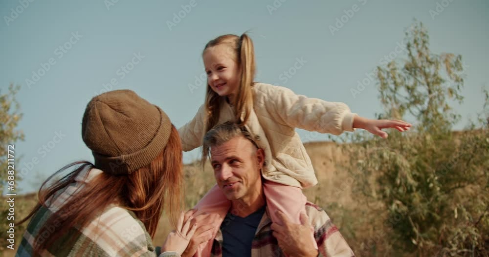 A little blonde girl has fun when she sits on the shoulders of her dad, the brunette man with a little gray hair in a checkered shirt, and talks to her mom, the brunette girl in a green checkered A little blonde girl has fun when she sits on the shoulders of her dad, the brunette man with a little gray hair in a checkered shirt, and talks to her mom, the brunette girl in a green checkered
