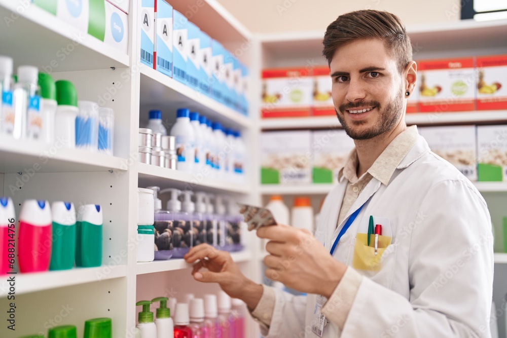 Young caucasian man pharmacist holding pills of shelving at pharmacy