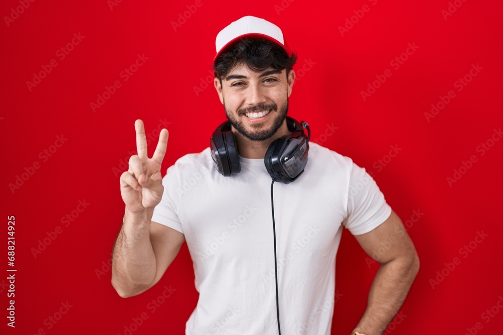 Hispanic man with beard wearing gamer hat and headphones showing and pointing up with fingers number two while smiling confident and happy. Hispanic man with beard wearing gamer hat and headphones showing and pointing up with fingers number two while smiling confident and happy.