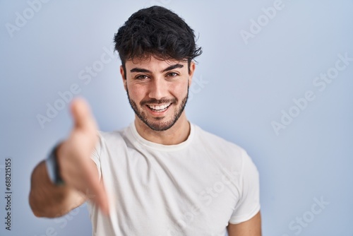 Hispanic man with beard standing over white background smiling friendly offering handshake as greeting and welcoming. successful business.