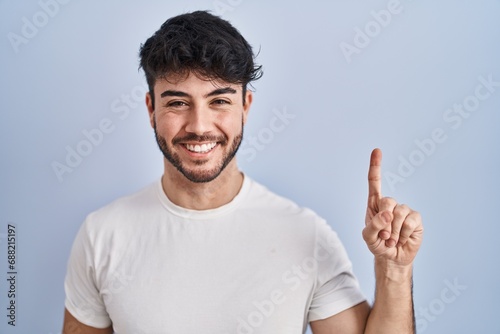 Hispanic man with beard standing over white background showing and pointing up with finger number one while smiling confident and happy. © Krakenimages.com