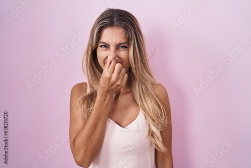 Young blonde woman standing over pink background smelling something stinky and disgusting, intolerable smell, holding breath with fingers on nose. bad smell