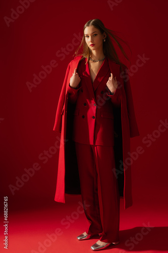 Fashionable confident woman wearing trendy red suit double breasted blazer, classic trousers, midi coat, metallic color shoes, posing on red background. Full-length studio fashion portrait
