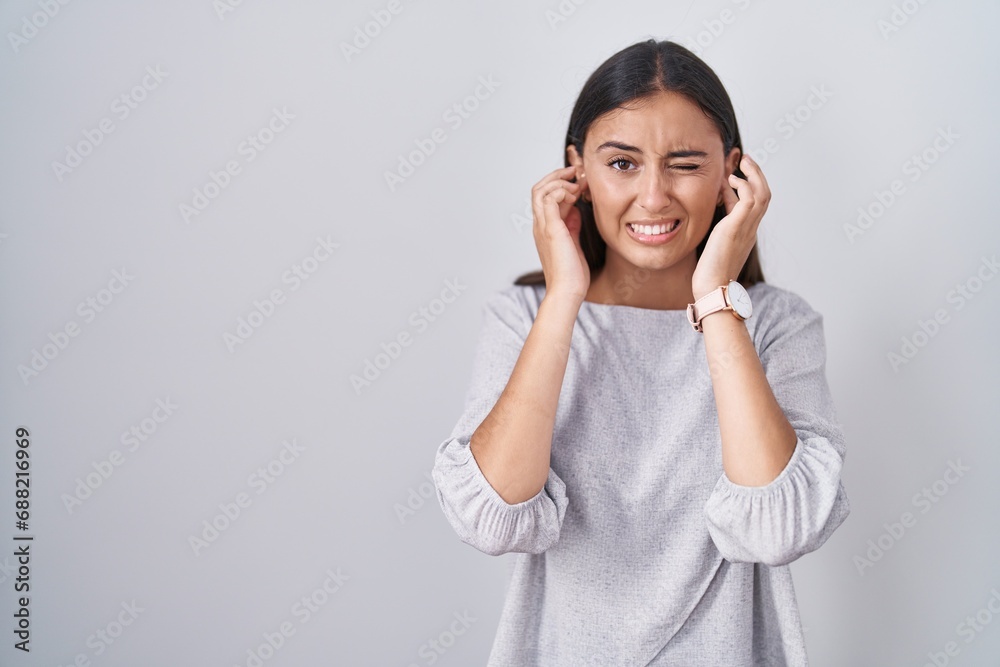 Young hispanic woman standing over white background covering ears with fingers with annoyed expression for the noise of loud music. deaf concept. Young hispanic woman standing over white background covering ears with fingers with annoyed expression for the noise of loud music. deaf concept.