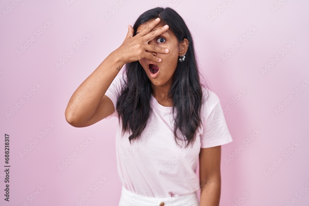 Young hispanic woman standing over pink background peeking in shock covering face and eyes with hand, looking through fingers with embarrassed expression. Young hispanic woman standing over pink background peeking in shock covering face and eyes with hand, looking through fingers with embarrassed expression.