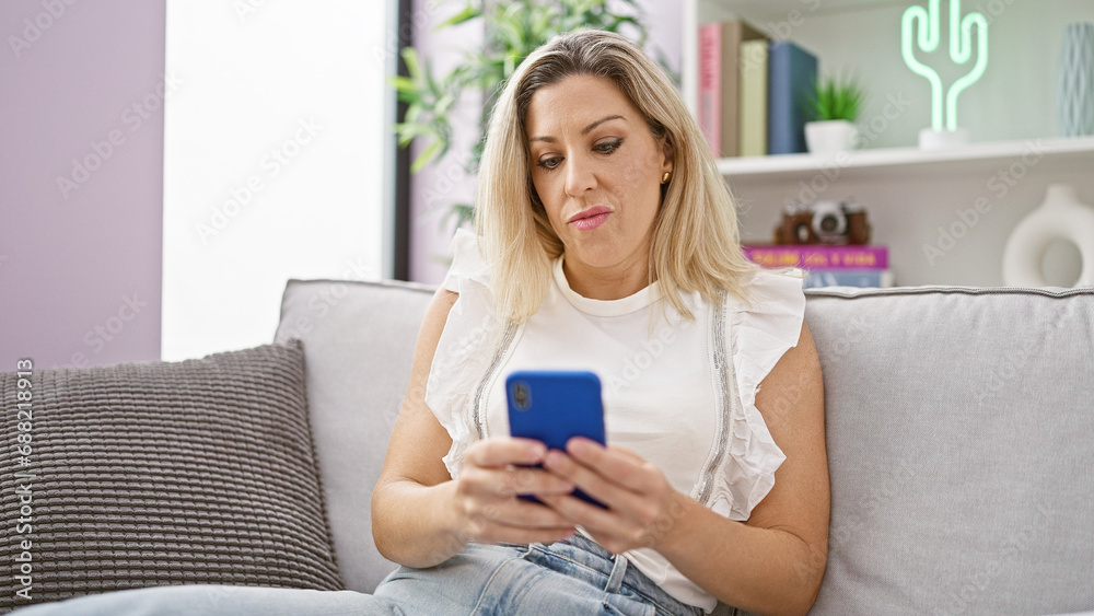 Young blonde woman using smartphone sitting on sofa with serious face at home Young blonde woman using smartphone sitting on sofa with serious face at home