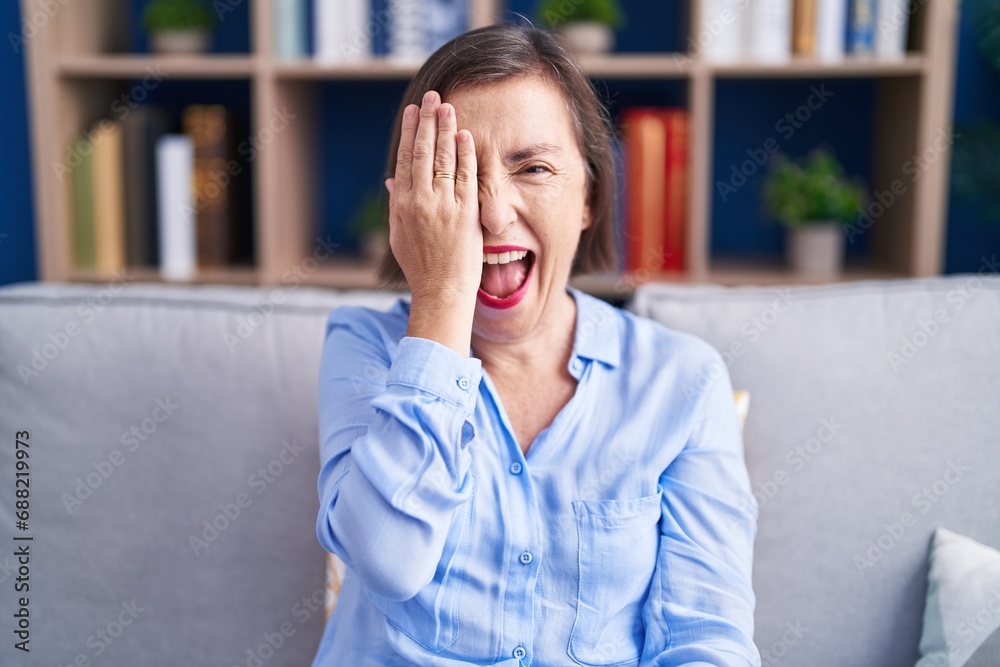 Middle age hispanic woman sitting on the sofa at home covering one eye with hand, confident smile on face and surprise emotion. Middle age hispanic woman sitting on the sofa at home covering one eye with hand, confident smile on face and surprise emotion.