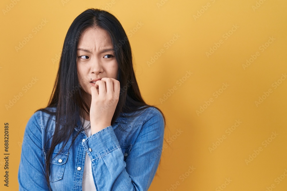 Young asian woman standing over yellow background looking stressed and nervous with hands on mouth biting nails. anxiety problem. Young asian woman standing over yellow background looking stressed and nervous with hands on mouth biting nails. anxiety problem.
