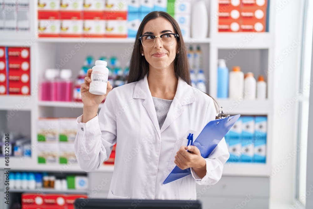 Young brunette woman working at pharmacy drugstore holding pills smiling looking to the side and staring away thinking.