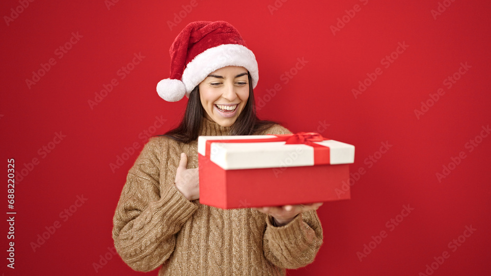 Young beautiful hispanic woman wearing christmas hat unpacking gift over isolated red background Young beautiful hispanic woman wearing christmas hat unpacking gift over isolated red background
