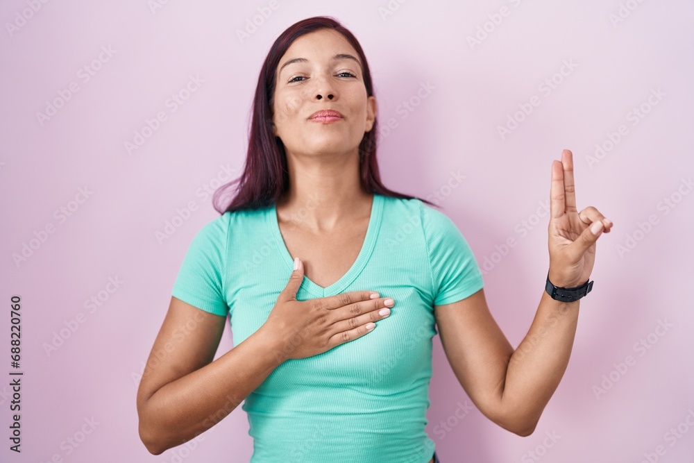 Young hispanic woman standing over pink background smiling swearing with hand on chest and fingers up, making a loyalty promise oath Young hispanic woman standing over pink background smiling swearing with hand on chest and fingers up, making a loyalty promise oath