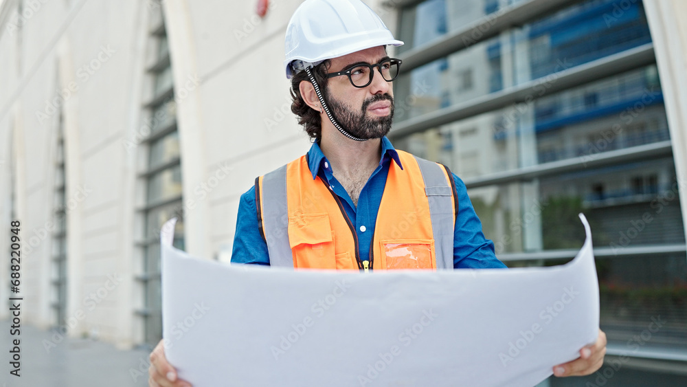 Young hispanic man architect holding blueprints looking around at construction place Young hispanic man architect holding blueprints looking around at construction place