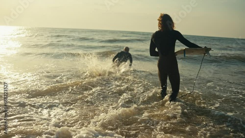Two men in wetsuits with surfboards go into the sea and ride the waves in summer. Two male surfers begin to swim in the sea at sunrise in summer