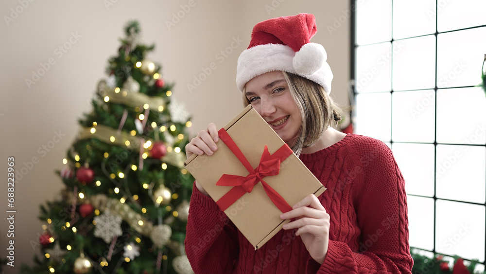 Young blonde woman holding gift standing by christmas tree at home Young blonde woman holding gift standing by christmas tree at home