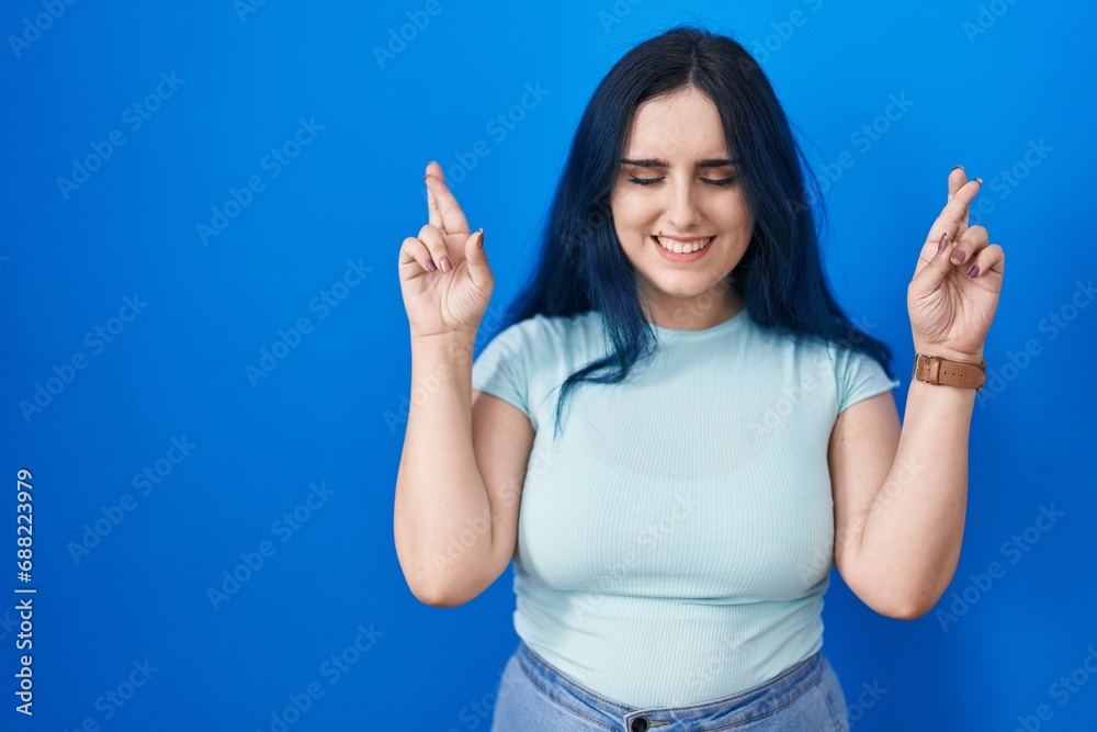Young modern girl with blue hair standing over blue background gesturing finger crossed smiling with hope and eyes closed. luck and superstitious concept. Young modern girl with blue hair standing over blue background gesturing finger crossed smiling with hope and eyes closed. luck and superstitious concept.