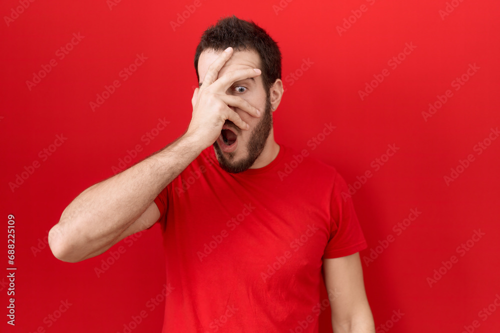 Young hispanic man wearing casual red t shirt peeking in shock covering face and eyes with hand, looking through fingers with embarrassed expression. Young hispanic man wearing casual red t shirt peeking in shock covering face and eyes with hand, looking through fingers with embarrassed expression.