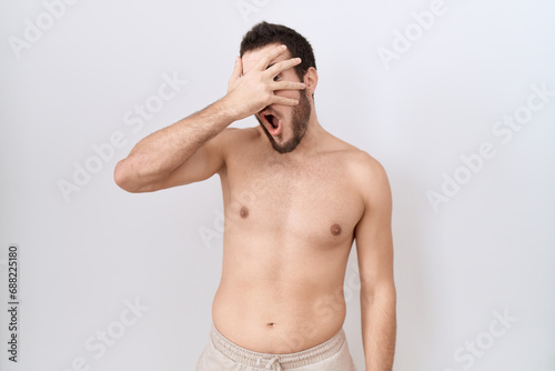 Young hispanic man standing shirtless over white background peeking in shock covering face and eyes with hand, looking through fingers with embarrassed expression.