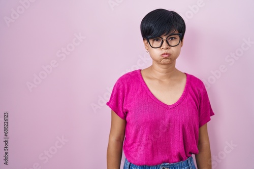 Young asian woman with short hair standing over pink background puffing cheeks with funny face. mouth inflated with air, crazy expression. photo