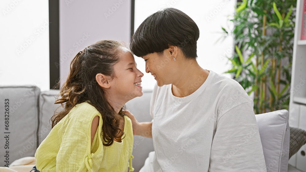 Mother and daughter enjoying a fun moment together at home, smiling confidently and touching noses, while comfortably sitting on living room sofa Mother and daughter enjoying a fun moment together at home, smiling confidently and touching noses, while comfortably sitting on living room sofa