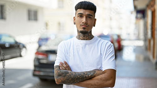 Cool young latin man with tattooed arms, standing outdoors on the urban street, carrying a relaxed yet serious expression with his arms casually crossed under the sunny city skyline.