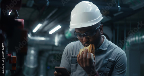 African American engineer eats sandwich and surfs the Internet on phone. Professional worker in uniform and protective hard hat having break working on industrial factory or energy facility. Close up.