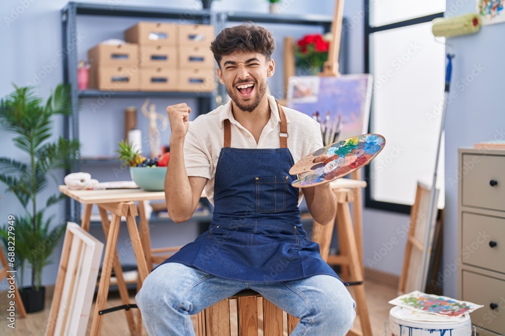 Arab man with beard painter sitting at art studio holding palette screaming proud, celebrating victory and success very excited with raised arms Arab man with beard painter sitting at art studio holding palette screaming proud, celebrating victory and success very excited with raised arms
