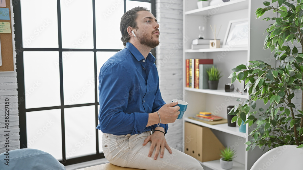 Young hispanic man business worker wearing earphones drinking coffee at the office