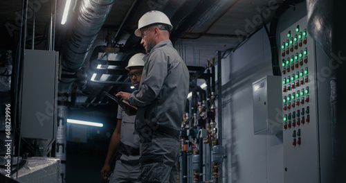 Professional Caucasian engineer in safety uniform, goggles and hard hat uses digital tablet computer, adjusts energy system on modern factory. Technician worker talks with African American colleague.