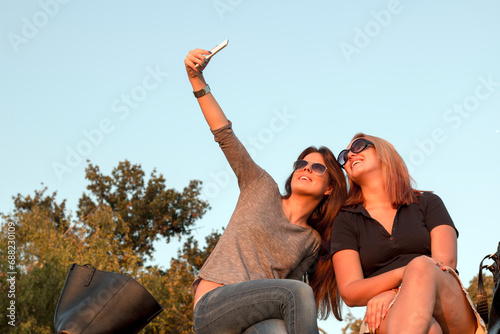 Two attractive young women with sunglasses sitting on the wall in public park and taking selfie photo. Time for selfie