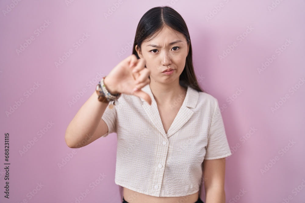 Chinese young woman standing over pink background looking unhappy and angry showing rejection and negative with thumbs down gesture. bad expression. Chinese young woman standing over pink background looking unhappy and angry showing rejection and negative with thumbs down gesture. bad expression.
