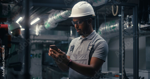 African American engineer uses mobile phone to inspect pipeline system on plant. Professional male worker in safety uniform and protective hard hat works on modern factory or industrial facility.