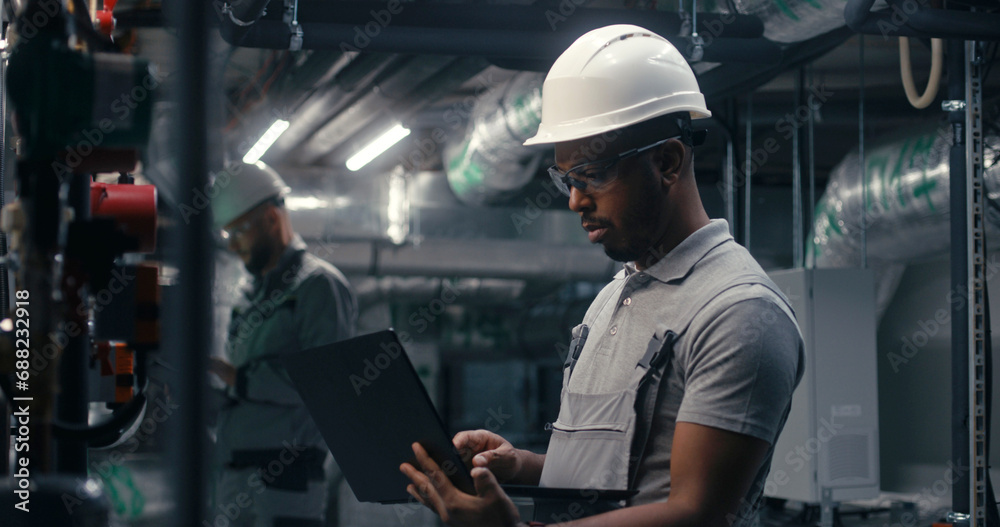 Professional African American inspector checks piping system on modern factory. Multiethnic team of technician specialists work on plant using laptop and tablet computers. Concept of heavy industry. Professional African American inspector checks piping system on modern factory. Multiethnic team of technician specialists work on plant using laptop and tablet computers. Concept of heavy industry.