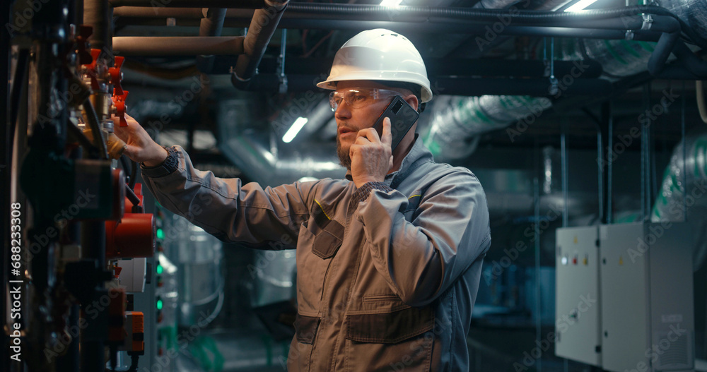 Male heavy industry worker wearing safety uniform, protective glasses and hard hat inspects pipeline system, talks by phone. Engineer maintains modern manufacturing factory or industrial facility. Male heavy industry worker wearing safety uniform, protective glasses and hard hat inspects pipeline system, talks by phone. Engineer maintains modern manufacturing factory or industrial facility.
