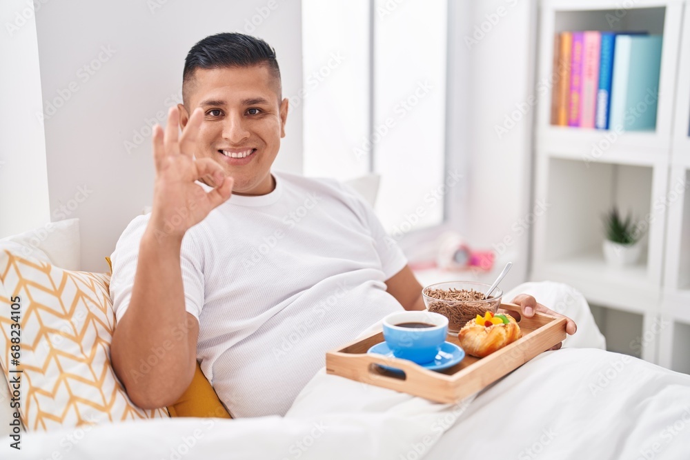 Young hispanic man eating breakfast in the bed doing ok sign with fingers, smiling friendly gesturing excellent symbol Young hispanic man eating breakfast in the bed doing ok sign with fingers, smiling friendly gesturing excellent symbol