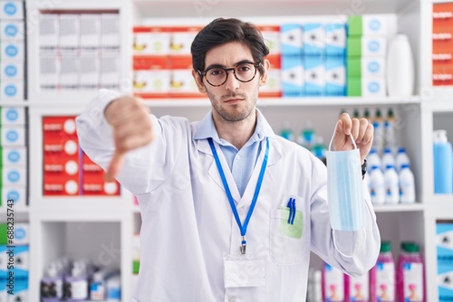 Young hispanic man working at pharmacy drugstore holding safety mask with angry face, negative sign showing dislike with thumbs down, rejection concept