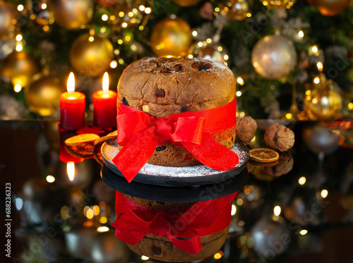 Italian Christmas cake Panettone with a red bow on a black glass against the background of a decorated Christmas tree