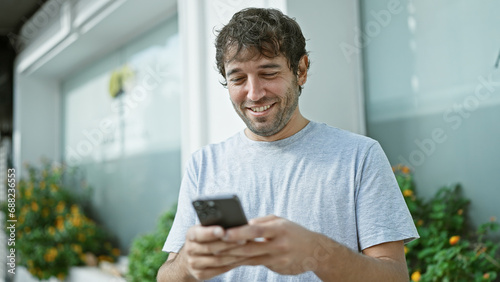 Happy young man, sporting a beard and blond hair, enjoying his time outdoor in the city, confidently smiling while typing a message on his smartphone screen