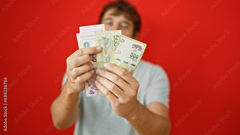 Young hispanic man, with a blond beard, caught in a portrait, counting his argentina pesos. standing isolated, engulfed in the rich red background, savings and business investment merge. Young hispanic man, with a blond beard, caught in a portrait, counting his argentina pesos. standing isolated, engulfed in the rich red background, savings and business investment merge.