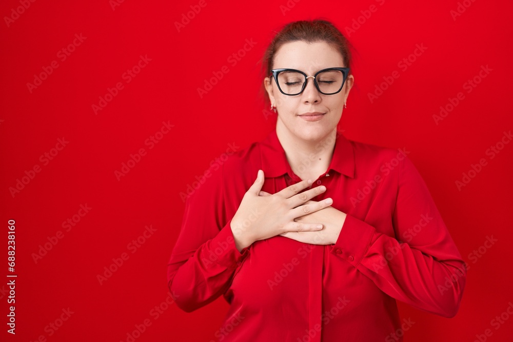 Young hispanic woman with red hair standing over red background smiling with hands on chest with closed eyes and grateful gesture on face. health concept. Young hispanic woman with red hair standing over red background smiling with hands on chest with closed eyes and grateful gesture on face. health concept.