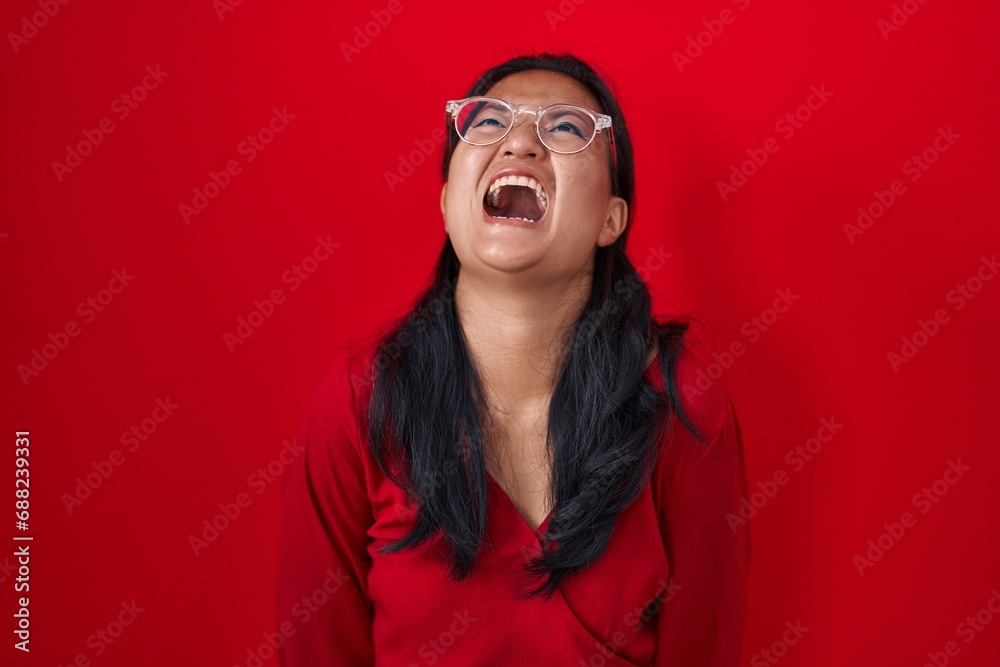 Asian young woman standing over red background angry and mad screaming frustrated and furious, shouting with anger. rage and aggressive concept.