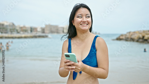 Young chinese woman tourist wearing bikini using smartphone at the beach