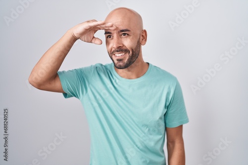 Middle age bald man standing over white background very happy and smiling looking far away with hand over head. searching concept.